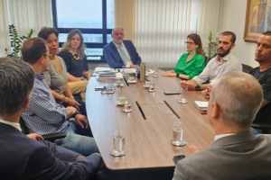 A imagem mostra uma reunião em uma sala com mesa retangular de madeira, com representantes do SINJUS e da 1ª Vice-Presidência do TJMG sentado ao redor da mesa. Todos estão atentos à discussão, com papéis e cadernos sobre a mesa.
