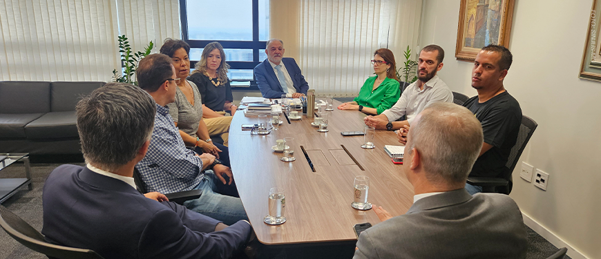 A imagem mostra uma reunião em uma sala com mesa retangular de madeira, com representantes do SINJUS e da 1ª Vice-Presidência do TJMG sentado ao redor da mesa. Todos estão atentos à discussão, com papéis e cadernos sobre a mesa.