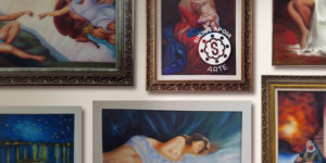 Imagem Acessível: A imagem mostra uma parede com várias pinturas emolduradas de estilo clássico e religioso, incluindo figuras humanas e cenas bíblicas. Em destaque no centro, o texto: “SINJUS Apoia: filiada aposentada retoma a arte e expõe suas obras em BH”. No canto superior direito, há o crédito “Foto: Rosana Marchetto” e o selo “SINJUS Apoia Arte”.