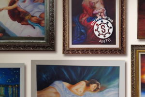 Imagem Acessível: A imagem mostra uma parede com várias pinturas emolduradas de estilo clássico e religioso, incluindo figuras humanas e cenas bíblicas. Em destaque no centro, o texto: “SINJUS Apoia: filiada aposentada retoma a arte e expõe suas obras em BH”. No canto superior direito, há o crédito “Foto: Rosana Marchetto” e o selo “SINJUS Apoia Arte”.