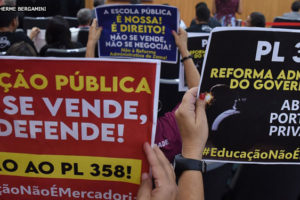 Servidores em primeiro plano segurando cartazes de protesto em um auditário lotado de pessoas contra o PL 358