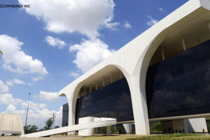 A imagem mostra a Cidade Administrativa de Minas Gerais, sede do governo estadual. O edifício na cor branca é moderno, com arquitetura contemporânea e apresenta uma fachada com grandes janelas que refletem o céu azul e as nuvens. Há uma grande abóbada sobre a entrada principal. Ao lado do edifício, há palmeiras. Ao fundo, o céu está parcialmente nublado.