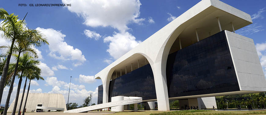 A imagem mostra a Cidade Administrativa de Minas Gerais, sede do governo estadual. O edifício na cor branca é moderno, com arquitetura contemporânea e apresenta uma fachada com grandes janelas que refletem o céu azul e as nuvens. Há uma grande abóbada sobre a entrada principal. Ao lado do edifício, há palmeiras. Ao fundo, o céu está parcialmente nublado.