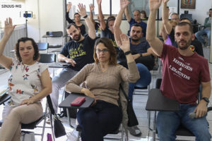 Grupo de pessoas reunidas, elas estão sentadas em cadeiras enfileiradas e exibem as mãos para o alto em sinal de aprovação às propostas apresentadas durante a Assembleia. Conteúdo textual: DELIBERAÇÃO - AGE AUTORIZA USO DO FUNDO DE GREVE NA LUTA CONTRA O RRF.