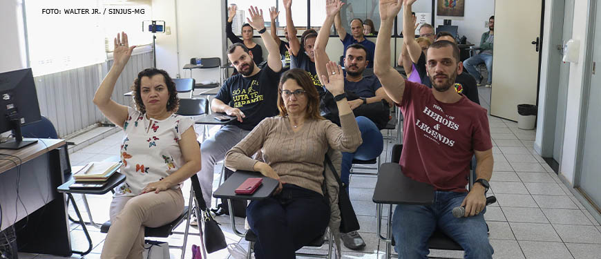 Grupo de pessoas reunidas, elas estão sentadas em cadeiras enfileiradas e exibem as mãos para o alto em sinal de aprovação às propostas apresentadas durante a Assembleia. Conteúdo textual: DELIBERAÇÃO - AGE AUTORIZA USO DO FUNDO DE GREVE NA LUTA CONTRA O RRF.