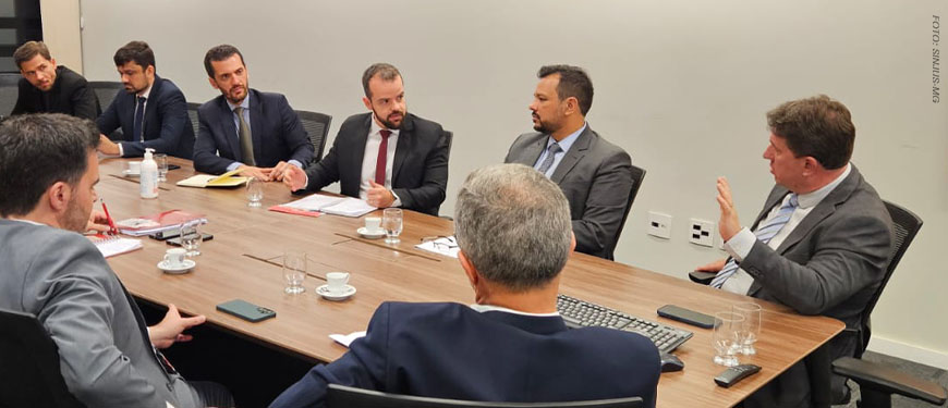 A imagem mostra uma reunião institucional, com homens de terno sentados ao redor de uma mesa. Entre os participantes estão representantes da presidência do Tribunal de Justiça de Minas Gerais, o coordenador-geral do SINJUS-MG, Felipe Rodrigues, e o diretor administrativo do Sindicato, Alexandre Pires. O sindicalista Felipe Rodrigues gesticula energicamente, enquanto dirige o olhar para o juiz auxiliar Thiago Colnago.