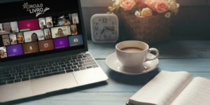 Imagem Acessível: A imagem mostra um ambiente aconchegante com uma mesa de madeira azul, um notebook exibindo uma videoconferência com várias pessoas e o logotipo “Círculo do Livro”, além de uma xícara de café, um livro aberto e um vaso com flores. Na parte inferior, o texto diz: “Reflexão sobre masculinidade saudável marca o início da nova temporada do Círculo do Livro do NM”.