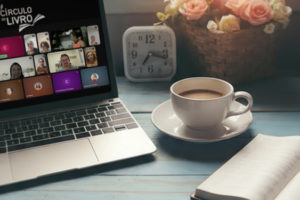 Imagem Acessível: A imagem mostra um ambiente aconchegante com uma mesa de madeira azul, um notebook exibindo uma videoconferência com várias pessoas e o logotipo “Círculo do Livro”, além de uma xícara de café, um livro aberto e um vaso com flores. Na parte inferior, o texto diz: “Reflexão sobre masculinidade saudável marca o início da nova temporada do Círculo do Livro do NM”.
