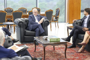 A imagem mostra um grupo de magistrados e autoridades reunidos em uma sala ampla e elegante, com poltronas de couro, mesa de centro e tapete vermelho. Ao fundo, grandes janelas revelam uma paisagem verde.
