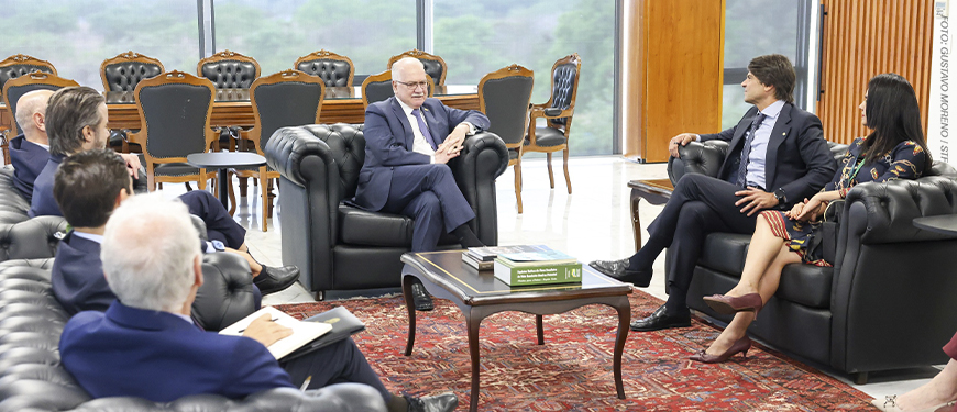 A imagem mostra um grupo de magistrados e autoridades reunidos em uma sala ampla e elegante, com poltronas de couro, mesa de centro e tapete vermelho. Ao fundo, grandes janelas revelam uma paisagem verde.
