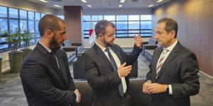 Dois dirigentes do SINJUS-MG conversam em uma sala ampla do Tribunal de Justiça de Minas Gerais (TJMG). Eles estão de terno e gravata, em pé, em frente a grandes janelas com vista para a cidade.