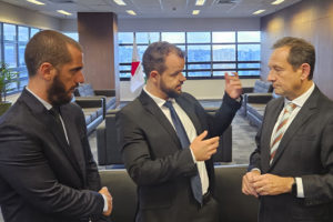 Dois dirigentes do SINJUS-MG conversam em uma sala ampla do Tribunal de Justiça de Minas Gerais (TJMG). Eles estão de terno e gravata, em pé, em frente a grandes janelas com vista para a cidade.