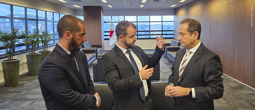 Dois dirigentes do SINJUS-MG conversam em uma sala ampla do Tribunal de Justiça de Minas Gerais (TJMG). Eles estão de terno e gravata, em pé, em frente a grandes janelas com vista para a cidade.