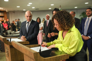 Edilene Lobo (mulher negra de cabelos cacheados castanhos de comprimento médio) está em um púlpito do Tribunal Superior Eleitoral, assinando o seu documento de posse, ela veste um traje social, em um púlpito ao lado dela está Alexandre de Moraes, um homem calvo, vestido em traje social azul marinho.