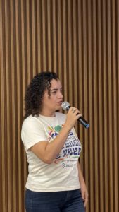 Adriana Teodoro, mulher branca de cabelos cacheados, fala ao microfone.