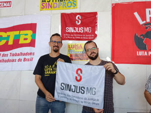 Felipe Rodrigues, o diretor de Assuntos Jurídicos, e ao seu lado está Alexandre Gomes, diretor de Comunicação e Imprensa do SINJUS-MG. Ambos estão de pé, segurando uma bandeira cinza com o logo do Sindicato. Atras, em uma parede branca, estão afixadas outras bandeiras e cartazes de entidades sindicais 