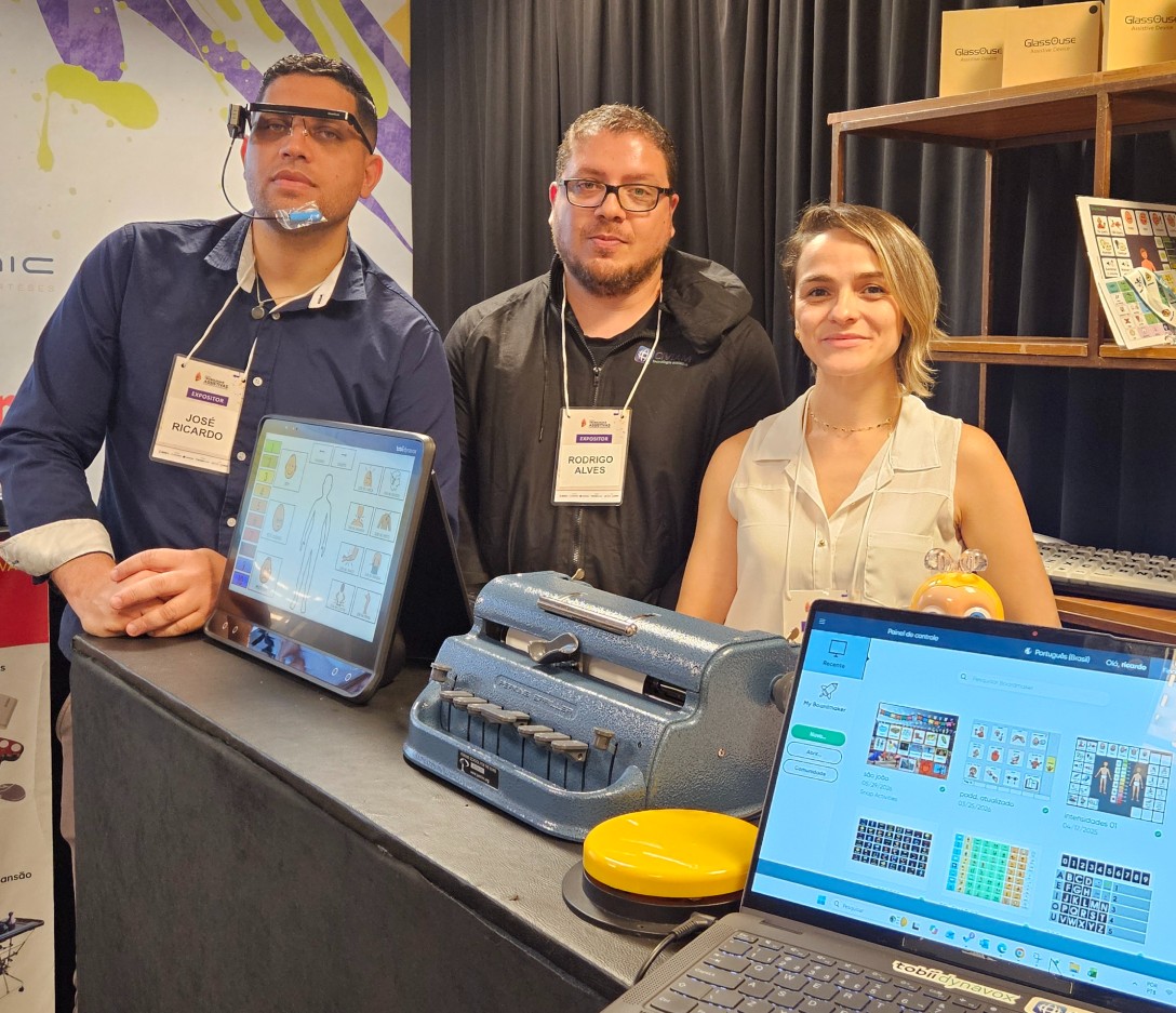 Fotografia registra o estande da Civiam Tecnologia Assistiva na Mostra de Tecnologias Assistivas e Soluções de Acessibilidade, promovida pelo SINJUS-MG. Da esquerda para a direita estão José Ricardo, Rodrigo Alves e Daiane Carvalho, posicionados atrás de uma bancada com diferentes recursos de tecnologia assistiva. José Ricardo utiliza um dispositivo eletrônico preso à cabeça, com estrutura semelhante a óculos e um pequeno componente próximo ao queixo. À frente dele há um tablet exibindo uma interface com símbolos e figuras para comunicação. No centro da bancada está uma máquina de escrever em braile, de estrutura metálica azul. Em primeiro plano, à direita, há um notebook aberto com uma plataforma de comunicação alternativa e aumentativa, com pranchas de símbolos, letras e outros recursos visuais. Ao lado do computador, aparece um grande acionador circular amarelo. Ao fundo, prateleiras exibem outros equipamentos e caixas de produtos de tecnologia assistiva.