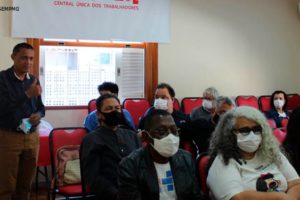 Homem negro com cabelos curtos e castanhos está vestido com camisa azul de manga longa e calça bege (Jonas Pinheiro, diretor de formação e política sindical). Ele discursa para diversas pessoas em um auditório com diversos ouvintes. Nas paredes há uma bandeira da CUT - Central Única dos Trabalhadores. Conteúdo textual: Mobilização - SINJUS e entidades sindicais se organizam para intensificar luta contra o RRF em MG.