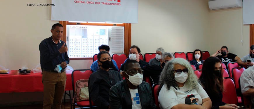 Homem negro com cabelos curtos e castanhos está vestido com camisa azul de manga longa e calça bege (Jonas Pinheiro, diretor de formação e política sindical). Ele discursa para diversas pessoas em um auditório com diversos ouvintes. Nas paredes há uma bandeira da CUT - Central Única dos Trabalhadores. Conteúdo textual: Mobilização - SINJUS e entidades sindicais se organizam para intensificar luta contra o RRF em MG.