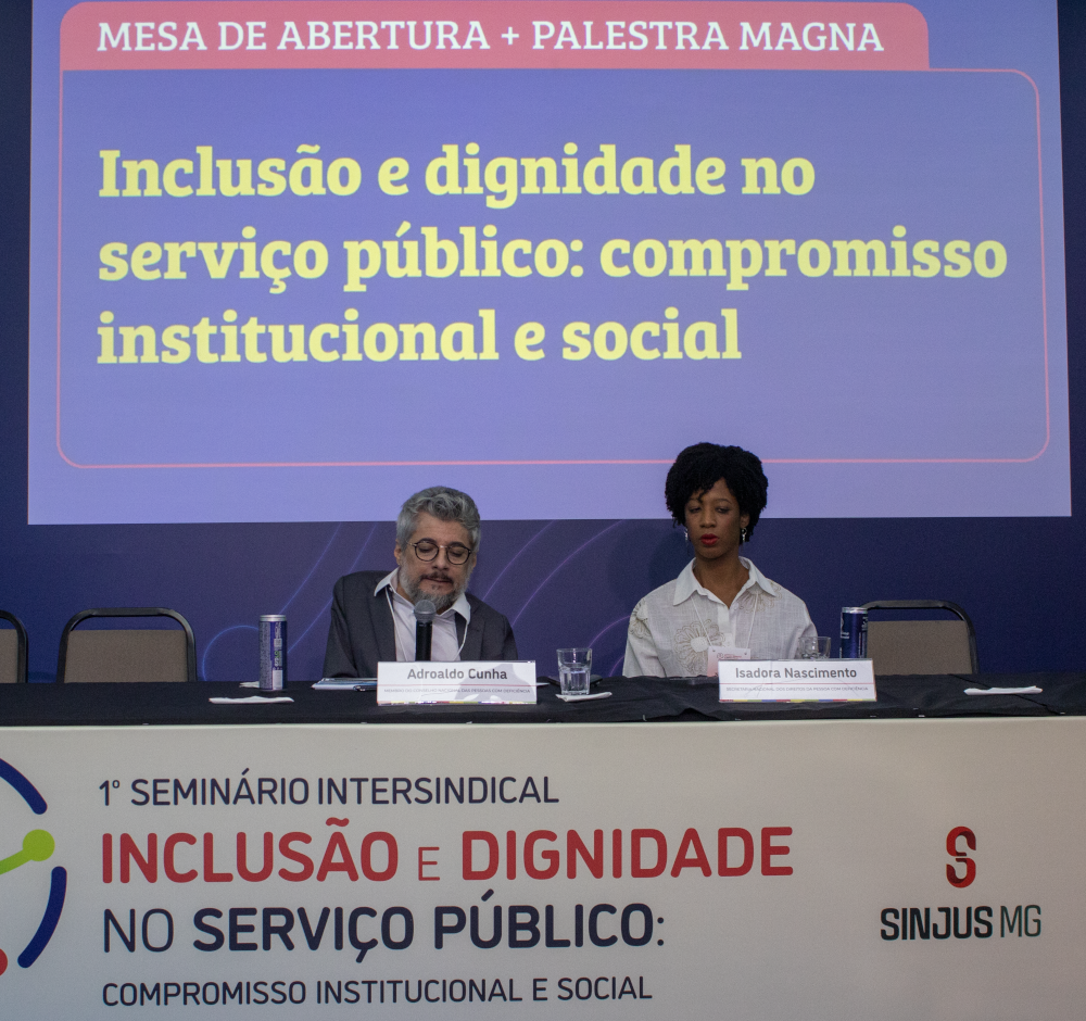 Mesa de abertura do 1º Seminário Intersindical de Inclusão e Dignidade no Serviço Público, promovido pelo SINJUS-MG. À esquerda, Adroaldo Portal, homem branco de cabelos grisalhos e óculos, fala ao microfone. À direita, Isadora Nascimento, mulher negra de cabelos crespos presos, veste camisa branca e olha com atenção. Ao fundo, telão com o título “Inclusão e dignidade no serviço público: compromisso institucional e social”. Na frente da mesa, painel com o nome do evento e logomarca do SINJUS-MG.