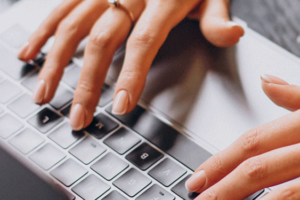 A imagem mostra as duas mãos de uma mulher digitando um notebook. Ao lado há uma xícara e um pires