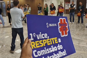 Em primeiro plano há uma pessoa segurando um cartaz com a mensagem "1º vice, respeite a Comissão do Teletrabalho", ao fundo, estão diversos servidores que participaram do ato público no saguão da sede do TJMG.