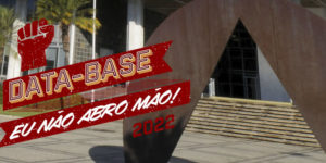 A imagem mostra o prédio da Assembleia Legislativa de Minas Gerais com uma escultura do artista Amilcar de Castro em primeiro plano. A escultura é um semi circulo em metal marrom com um um corte central triangular. Ao fundo do prédio o céu é azul.