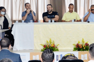 m grupo de 7 pessoas reunidas em frente a uma plateia e em uma mesa decorada com tecido amarelo e 3 arranjos de flores. Destaque para o coordenador-geral do SINJUS-MG, Alexandre Pires, que está falando ao microfone. Ao fundo uma cortina de cor marrom, semitransparente. Ao todo aparecem 17 pessoas que estão na plateia aparecem na imagem. 