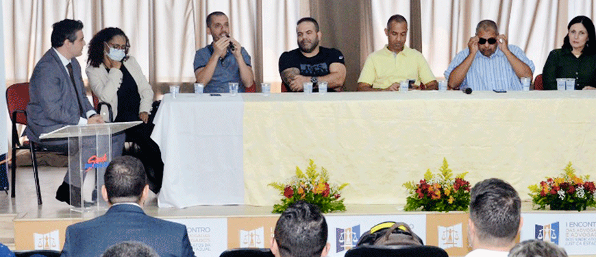 m grupo de 7 pessoas reunidas em frente a uma plateia e em uma mesa decorada com tecido amarelo e 3 arranjos de flores. Destaque para o coordenador-geral do SINJUS-MG, Alexandre Pires, que está falando ao microfone. Ao fundo uma cortina de cor marrom, semitransparente. Ao todo aparecem 17 pessoas que estão na plateia aparecem na imagem. 