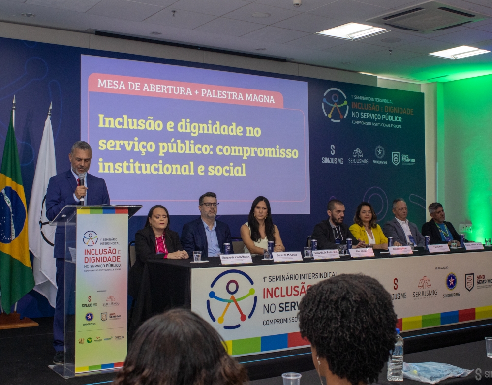 Em um auditório, ocorre a mesa de abertura do 1º Seminário Intersindical Inclusão e Dignidade no Serviço Público: Compromisso Institucional e Social. À esquerda da imagem, em pé atrás de um púlpito, está Alex Aguiar, diretor de Finanças e coordenador do Núcleo da Pessoa com Deficiência do SINJUS-MG. Ele é um homem de pele clara, cabelo curto grisalho e barba, veste terno azul e gravata vermelha, e lê ao microfone. Sentados à mesa, da esquerda para a direita: Simone Barros, servidora do TJMG, mulher de pele clara e cabelos castanhos escuros, veste blazer preto e blusa vermelha, com expressão atenta. Eduardo Couto, presidente do SERJUSMIG, homem de pele clara, óculos, cabelos curtos grisalhos e barba, veste camisa azul clara e blazer azul-escuro, olhando para a frente. Fernanda de Paula Silva, Promotora de Justiça de Minas Gerais, mulher de pele clara, cabelos lisos e longos, veste blusa branca sem mangas, com postura ereta e séria. Alexandre Pires, coordenador-geral do SINJUS, homem de pele clara, barba cheia, veste camiseta preta e blazer escuro, com semblante concentrado. Fanny Melo, coordenadora-geral do SINDSEMPMG, mulher de pele clara, cabelos castanhos médios, veste blazer cinza e blusa amarela, com expressão firme. Marcos do Vale, diretor-geral do SINDOJUS, homem de pele clara e cabelos grisalhos, veste camisa branca e blazer cinza, observando atentamente. Hércules Marcone, servidor do TJMG, homem de pele clara, cabelos curtos escuros, veste camiseta preta e blazer escuro, sentado à direita.