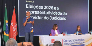 Painel do 13º Congresso dos Servidores da Justiça de Minas Gerais com o tema 'Eleições 2026 e a representatividade do servidor do Judiciário'. Um homem fala ao microfone em pé, enquanto três pessoas estão sentadas à mesa, em frente a um telão com o título do painel. Ao lado, estão expostas bandeiras do Brasil, de Minas Gerais e do SINJUS-MG.