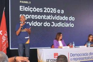 Painel do 13º Congresso dos Servidores da Justiça de Minas Gerais com o tema 'Eleições 2026 e a representatividade do servidor do Judiciário'. Um homem fala ao microfone em pé, enquanto três pessoas estão sentadas à mesa, em frente a um telão com o título do painel. Ao lado, estão expostas bandeiras do Brasil, de Minas Gerais e do SINJUS-MG.
