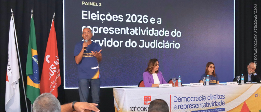 Painel do 13º Congresso dos Servidores da Justiça de Minas Gerais com o tema 'Eleições 2026 e a representatividade do servidor do Judiciário'. Um homem fala ao microfone em pé, enquanto três pessoas estão sentadas à mesa, em frente a um telão com o título do painel. Ao lado, estão expostas bandeiras do Brasil, de Minas Gerais e do SINJUS-MG.