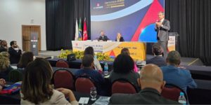Auditório com público assistindo a uma palestra durante o 13º Consinjus. Um homem de terno fala em pé ao lado do púlpito, enquanto duas pessoas estão sentadas à mesa de mediação no palco, em frente a um painel colorido com a frase ‘Democracia, Direitos e Representatividade’. Na mesa também há arranjos de flores amarelas e bandeiras do Brasil, de Minas Gerais e do SINJUS."