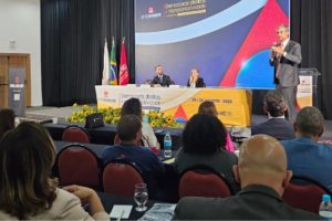 Auditório com público assistindo a uma palestra durante o 13º Consinjus. Um homem de terno fala em pé ao lado do púlpito, enquanto duas pessoas estão sentadas à mesa de mediação no palco, em frente a um painel colorido com a frase ‘Democracia, Direitos e Representatividade’. Na mesa também há arranjos de flores amarelas e bandeiras do Brasil, de Minas Gerais e do SINJUS."
