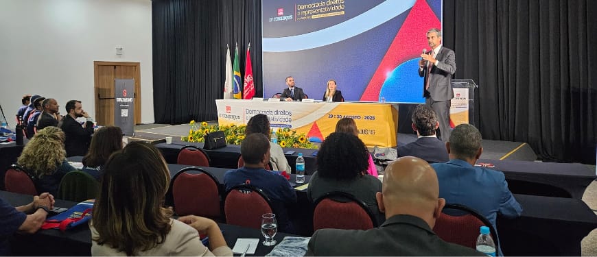 Auditório com público assistindo a uma palestra durante o 13º Consinjus. Um homem de terno fala em pé ao lado do púlpito, enquanto duas pessoas estão sentadas à mesa de mediação no palco, em frente a um painel colorido com a frase ‘Democracia, Direitos e Representatividade’. Na mesa também há arranjos de flores amarelas e bandeiras do Brasil, de Minas Gerais e do SINJUS."