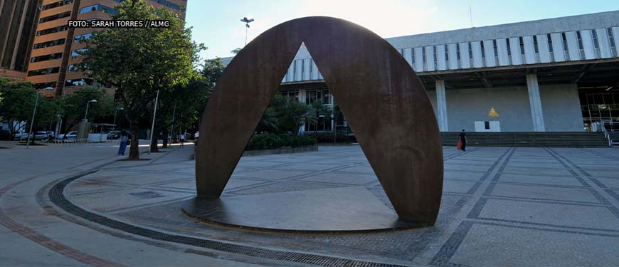 fachada da Assembleia Legislativa de Minas Gerais com destaque para o monumento que consiste em um triângulo vazado em um círculo de bronze.