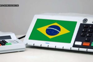fundo branco e em destaque urna eletrônica eleitoral com a bandeira do Brasil na tela.