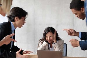 sala de reunião em que há uma mulher sentada em uma mesa com as mãos tampando os ouvidos e com expressão de sofrimento sentada. Em volta, homens e mulheres apontam o dedo para a referida mulher com expressões faciais hostis.