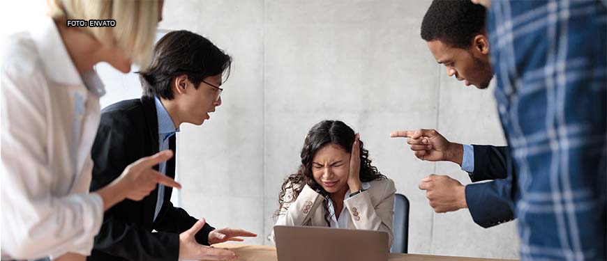 sala de reunião em que há uma mulher sentada em uma mesa com as mãos tampando os ouvidos e com expressão de sofrimento sentada. Em volta, homens e mulheres apontam o dedo para a referida mulher com expressões faciais hostis.