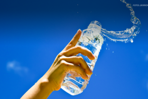 Imagem com um fundo de céu azul claro, onde uma mão levanta uma garrafa de água transparente, com parte da água sendo derramada.