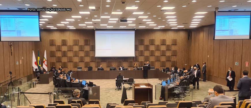 Imagem aberta do auditório do TJMG enquadrando as desembargadoras e os desembargadores membros do Órgão que estão sentados em formação semicircular, lado a lado. As paredes e o carpete do local são em tom marrom e os magistrados usam togas na cor preta.