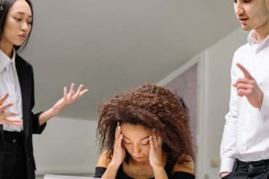 sala de reunião em que há uma mulher sentada em uma mesa com as mãos tampando os ouvidos e com expressão de sofrimento sentada. Em volta, homens e mulheres apontam o dedo para a referida mulher com expressões faciais hostis.