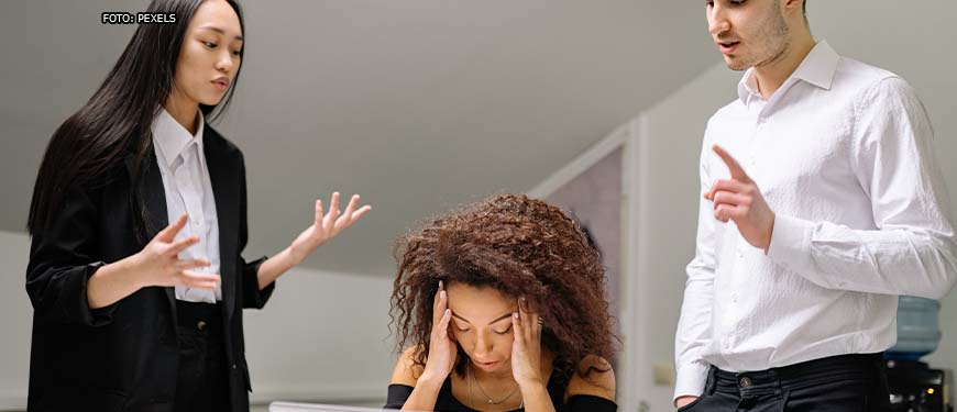 sala de reunião em que há uma mulher sentada em uma mesa com as mãos tampando os ouvidos e com expressão de sofrimento sentada. Em volta, homens e mulheres apontam o dedo para a referida mulher com expressões faciais hostis.