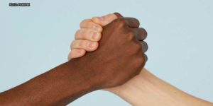 Enquadramento fechado mostrando duas mãos dadas, uma de pessoa de pele clara e outra de pessoa negra.
