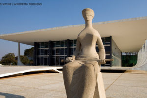 Foto da escultura "A Justiça", localizada em frente ao prédio do Supremo Tribunal Federal, na Praça dos Três Poderes, em Brasília. O monumento representa uma mulher, sentada e vendada, segurando uma espada, um dos símbolos mais usados para caracterizar as deusas greco-romanas das leis e da justiça. Conteúdo textual: Nota. Fenajud repudia ataque de Roberto Jefferson a policiais federais e afronta à decisão judicial.