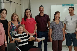 Foto do grupo que esteve presente no encontro do NAP, estão na foto, a partir da esquerda: uma mulher de pele clara, cabelos curtos e castanhos, vestindo uma blusa cor de rosa; uma mulher de pele clara e cabelos bem curtos, vestindo uma blusa preta; mulher de pele clara, loira, com uma blusa de alças finas bem colorida; uma mulher de cabelos médios e pele clara, vestindo uma blusa de cor vermelha; um homem alto, de pele clara com cabelos curtos e pouco grisalhos, vestindo uma blusa na cor vinho; uma mulher de pele clara e cabelos médios presos para trás, vestindo uma blusa nas cores preto e branco; um homem de pele parda e cabelos castanhos, vestindo uma blusa branca com um óculos de grau pendurado na gola; uma mulher de cabelos médios e castanhos, pele clara, vestindo uma blusa na cor preta. À frente de todos temos, sentada, uma mulher de pele clara, cabelos curtos e loiros, vestindo uma blusa listrada nas cores preto e branco. Conteúdo textual: APOSENTADOS E PENSIONISTAS. QUIZ DO MÊS DO SERVIDOR É ATRAÇÃO DO ENCONTRO DO NAP