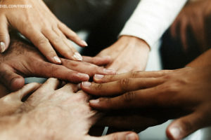 Foto de um grupo diverso de pessoas encostando suas mãos no centro de uma roda em sinal de união.