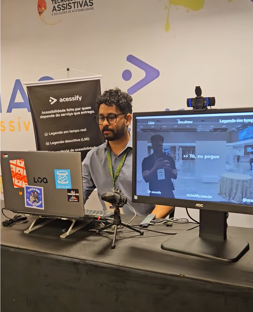 Matheus Guifer está no estande da Acessify durante a Mostra de Tecnologias Assistivas e Soluções de Acessibilidade, promovida pelo SINJUS-MG. Ele aparece atrás de uma bancada com notebook, câmera e monitor, demonstrando uma solução de acessibilidade. Na tela maior, é exibida uma legenda em tempo real sobre a imagem captada pela câmera. Ao fundo, um painel da Acessify apresenta informações sobre serviços como legenda em tempo real e legenda descritiva.