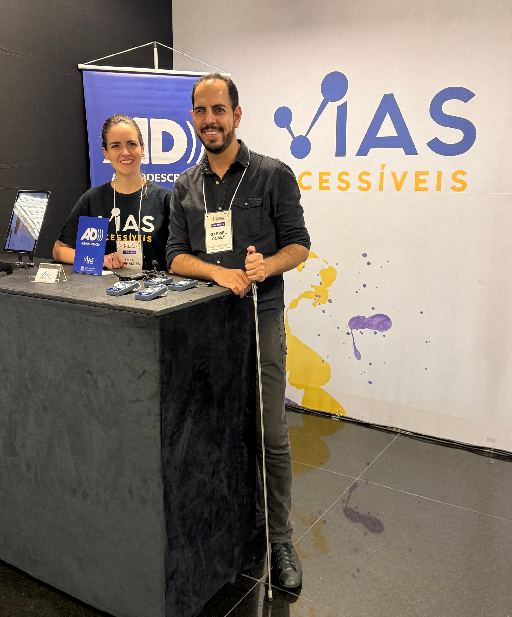 idia Fernandes e Gabriel Aquino posam no estande da Vias Acessíveis durante a Mostra de Tecnologias Assistivas e Soluções de Acessibilidade, promovida pelo SINJUS-MG. Lidia está atrás de uma bancada preta, usando camiseta da empresa e crachá, ao lado de materiais de divulgação e equipamentos de audiodescrição. Gabriel está à frente da bancada, também com crachá, vestindo camisa preta e segurando uma bengala branca. Ao fundo, um grande painel exibe a marca “Vias Acessíveis”, em azul e amarelo.
